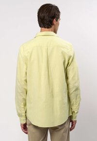 Mann trägt ein hellgelbes langärmeliges Hemd und beige Hosen, steht mit dem Rücken zur Kamera vor einem schlichten hellen Hintergrund.