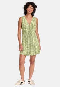 Grön gingham mini-klänning med V-ringning, knapplist fram och ärmlös design. Kombinerad med beige sneakers. Slät tygtextur.