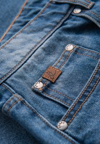 Blå denimjeanslomme med orange syning, metalnitter og et lille brunt lædermærke med et kursivt bogstav "R".