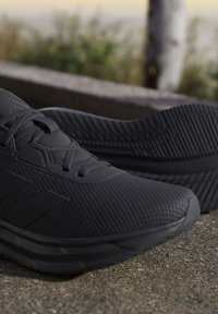 Paire de chaussures de sport noires avec empeigne en mesh texturé et semelle à motifs reposant sur une surface rugueuse en béton à l'extérieur.