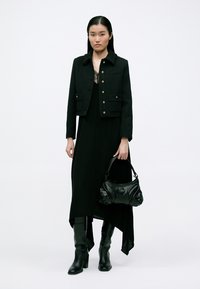 Giacca corta in tweed nero con dettagli in bottone dorato, indossata sopra un lungo vestito nero. La modella porta una borsa nera testurizzata e indossa stivali al ginocchio.