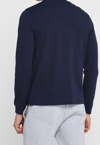 Námorně modrá košile s dlouhým rukávem s texturovaným povrchem, kulatým výstřihem a rovnými lemy, nošena se šedými tepláky.