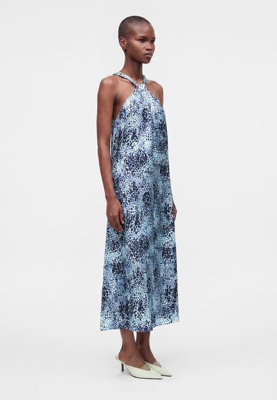 Femme mince debout portant une robe midi sans manches à motif bleu à col halter avec des chaussures à talons pointues vert clair.