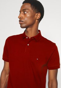 Polo rouge en tissu texturé. Doté d'un col, de deux boutons et d'un petit logo sur le côté gauche. Manches courtes.
