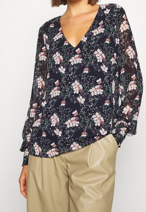 Blusa floreale navy con scollo a V profondo, maniche lunghe trasparenti e motivi floreali rosa/verdi, abbinata a pantaloni beige plissettati.