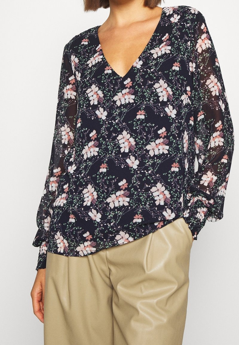 Granatowa bluzka w kwiaty z głębokim dekoltem w kształcie litery V, przezroczystymi długimi rękawami oraz różowo-zielonymi wzorami kwiatowymi, zestawiona z beżowymi spódnicami plisowanymi.