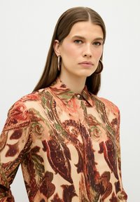 Blouse à col avec un motif floral tourbillonnant en brun, vert et corail sur un fond crème ; tissu doux avec une texture lisse.