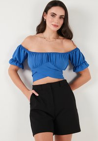 Blå off-the-shoulder top med puffede ærmer, struktureret stof og crossover-detalje ved taljen. Sammen med sorte skræddersyede shorts.
