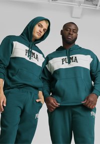 Due individui indossano felpe coordinate di colore blu teal con una striscia orizzontale bianca. Le felpe presentano un logo centrale "PUMA" in nero.