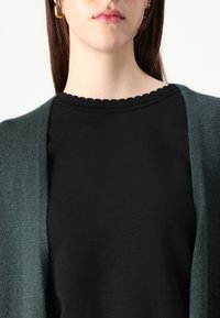 Tumma vihreä neuletakki, jossa on pehmeä tekstuuri, puettuna mustan paidan päälle, jossa on aaltoileva pääntie. Selkeä, yksinkertainen muotoilu ilman kuvioita.