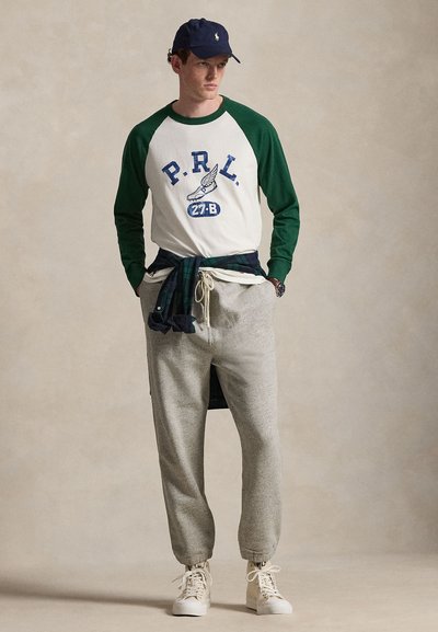 Νέος άνδρας με μακρυμάνικο πουκάμισο σε πράσινο και λευκό με το κείμενο "P.R.L.", γκρι φόρμα, λευκά αθλητικά παπούτσια, μπλε σκουφί και καρό πουκάμισο δεμένο στη μέση.
