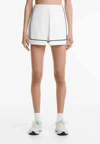 Hvide skræddersyede shorts med sort kant, glat stof, plisseret front og lige kant. Båret med hvide sneakers og sokker.