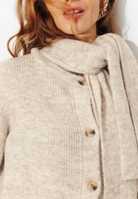 Gilet en maille beige avec un col revers et une longue écharpe, doté de boutons en bois et d'une texture douce. Accentué de détails côtelés.