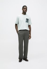 Camiseta de algodón gris claro con un bolsillo de estampado floral oscuro, combinada con pantalones gris oscuro. El modelo está de pie sobre un fondo neutro.