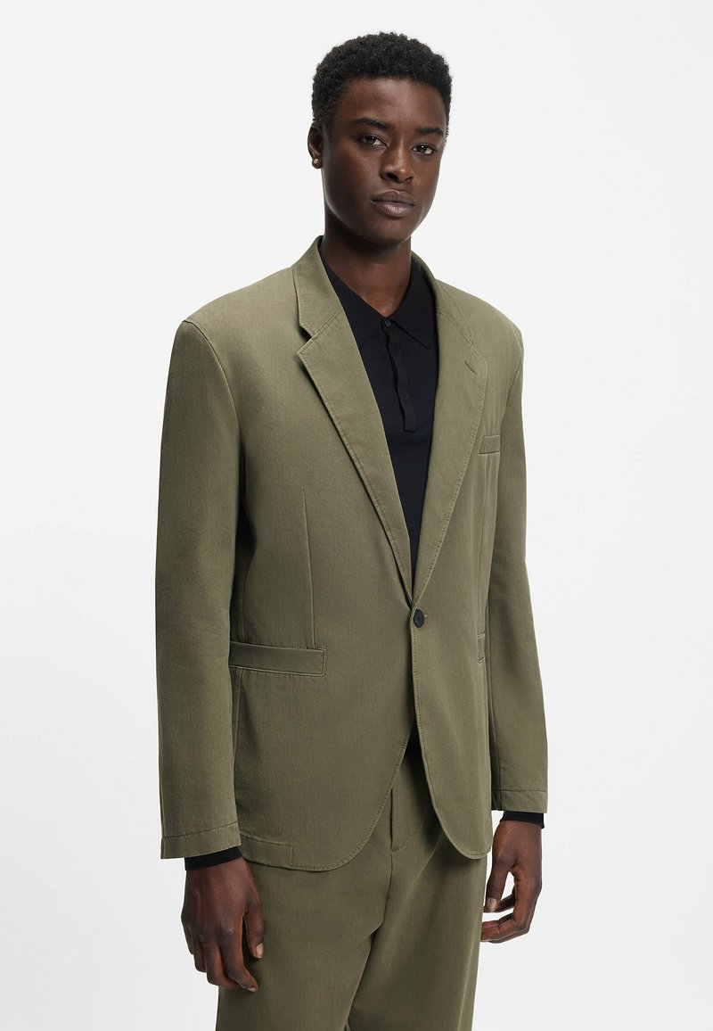 Homme portant une veste de costume vert olive avec un pantalon assorti sur une chemise noire à col, debout devant un fond blanc uni.