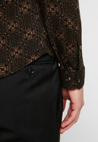 Camicia nera con motivo a fiori marroni, maniche lunghe e polsini con bottoni, abbinata a pantaloni neri con tasca laterale.