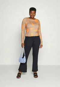 Mulher vestindo uma blusa de manga longa com padrão bege e laranja, calças pretas, sandálias pretas e segurando uma bolsa azul clara, em pé sobre um fundo simples.