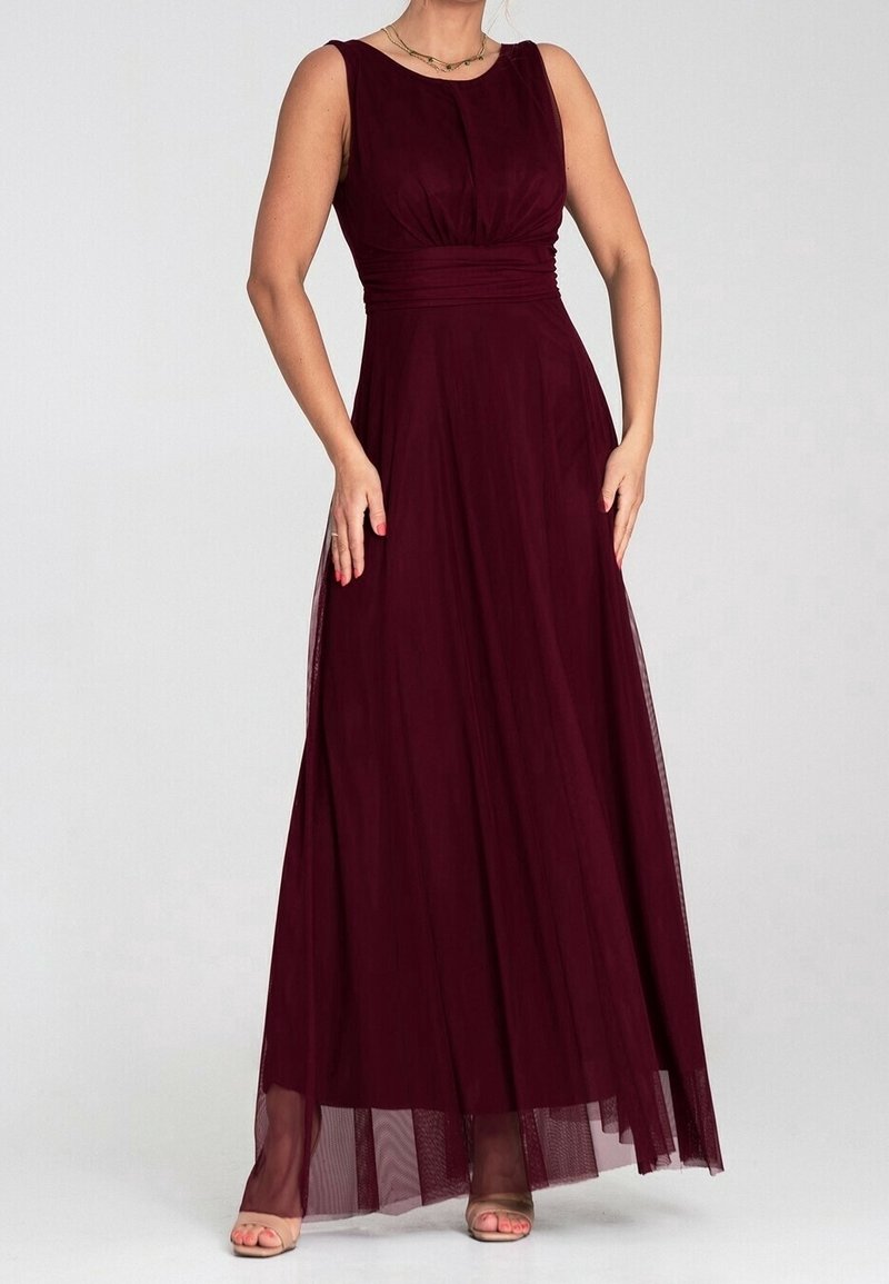 Femme portant une robe longue sans manches de couleur bordeaux foncé avec une taille froncée et une superposition transparente, debout devant un fond uni.