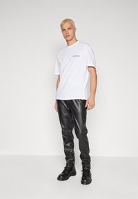 Weißes Baumwoll-T-Shirt mit kleinem schwarzem Logo, kombiniert mit schwarzen Kunstlederhosen und schwarzen Schnürstiefeln. Schlichter Hintergrund.