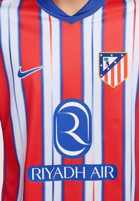 Punainen ja valkoinen raidoitettu jalkapallon pelipaita, jossa on sinisiä yksityiskohtia, Nike-logo, seuran vaakuna ja "RIYADH AIR" -brändäys edessä.