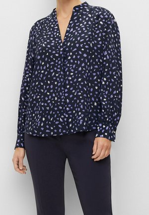 Femme portant un chemisier foncé avec des taches irrégulières lavande et blanches, manches longues et col en V sans col, associé à un pantalon foncé.