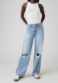 Světle modré džíny s širokými nohavicemi z denimu s opranými koleny a roztrhanými okraji, doplněné bílým ribovaným tílkem a teniskami.