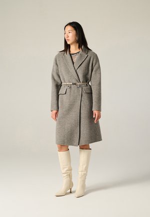Cappotto di lana grigio con vita regolabile, ampi rever e due tasche frontali, abbinato a stivali a stiletto color crema al ginocchio.