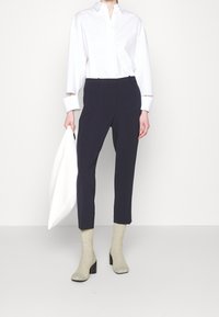 Camisa blanca de manga larga con botones, pantalones azul marino ajustados, calcetines de punto claro y zapatos negros de tacón ancho, sosteniendo un bolso blanco.