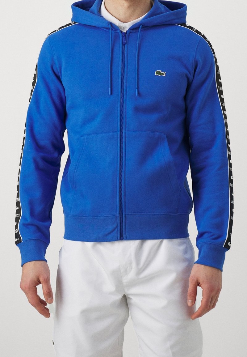 Homme portant un sweat à capuche zippé bleu vif avec des bandes noires et blanches sur les manches et un pantalon blanc sur fond uni.