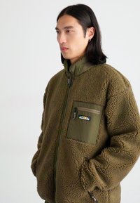 Brązowa polarowa kurtka z wysokim kołnierzem, zamek błyskawiczny i przednia kieszeń w kolorze oliwkowym. Posiada naszywkę z logo i fakturowany materiał.