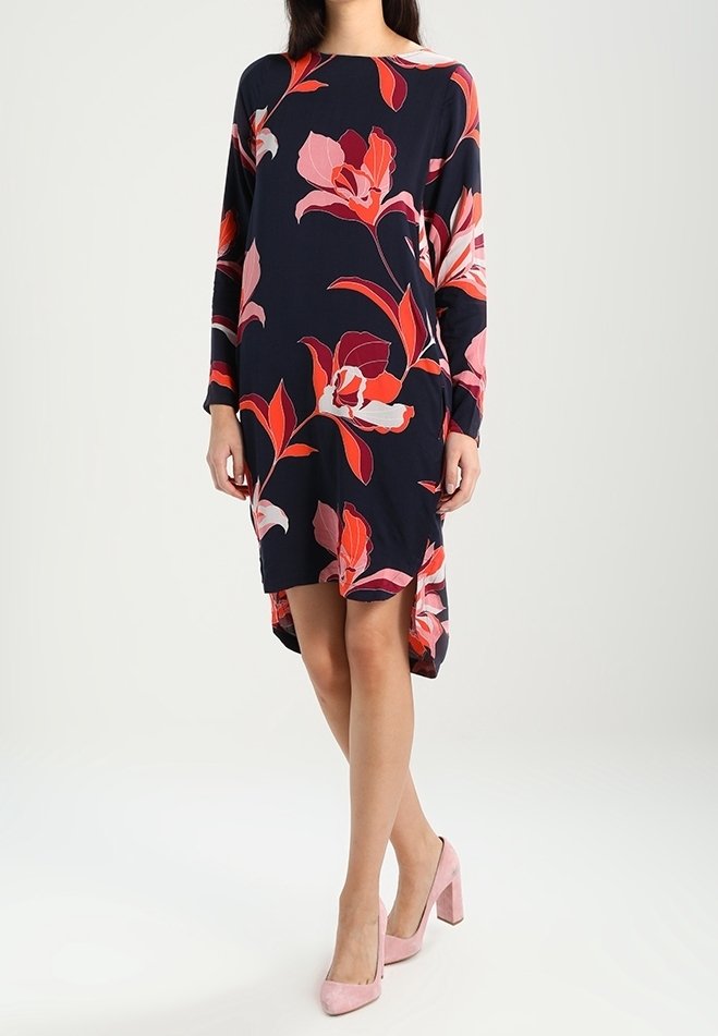 Femme portant une robe à manches longues bleu marine avec de grands motifs floraux roses et rouges, assortie à des chaussures à talons roses.