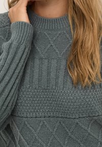 Primo piano di una persona che indossa un maglione grigio a maglia texturizzata con motivi a rombi e coste, con lunghi capelli biondi e una mano vicino al collo.