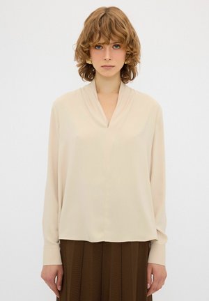 Beige Bluse mit V-Ausschnitt, langen Ärmeln und weicher Textur. Der Stoff wirkt leicht und fällt geschmeidig mit einer lockeren Passform.