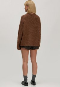 Frau mit kurzen blonden Haaren, die einen braunen, flauschigen Pullover, dunkle Shorts, graue Socken und schwarze Slipper trägt, steht vor einem schlichten Hintergrund.