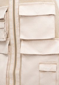 Μπεζ καπιτονέ γιλέκο με πολλαπλές μπροστινές τσέπες, διαθέτει κεντρικό φερμουάρ, ριμπ υφή και λεία υφή υφάσματος.