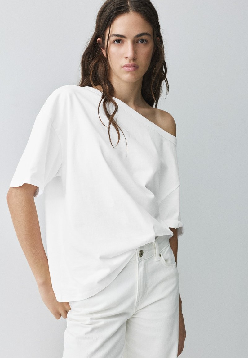 Jeune femme aux longs cheveux ondulés porte un t-shirt blanc à épaules dénudées rentré dans un pantalon blanc taille haute, debout devant un fond uni.