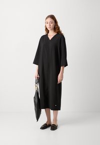 Robe noire, mi-longue, avec un décolleté en v, des manches trois-quarts et une coupe décontractée. Porte des mocassins noirs et tient un sac noir.