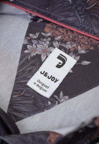 Paarse stof met bloemmotieven en grijze en oranje accenten. Zichtbaar label leest "J&JOY, Ontworpen in België." Textuurontwerp.