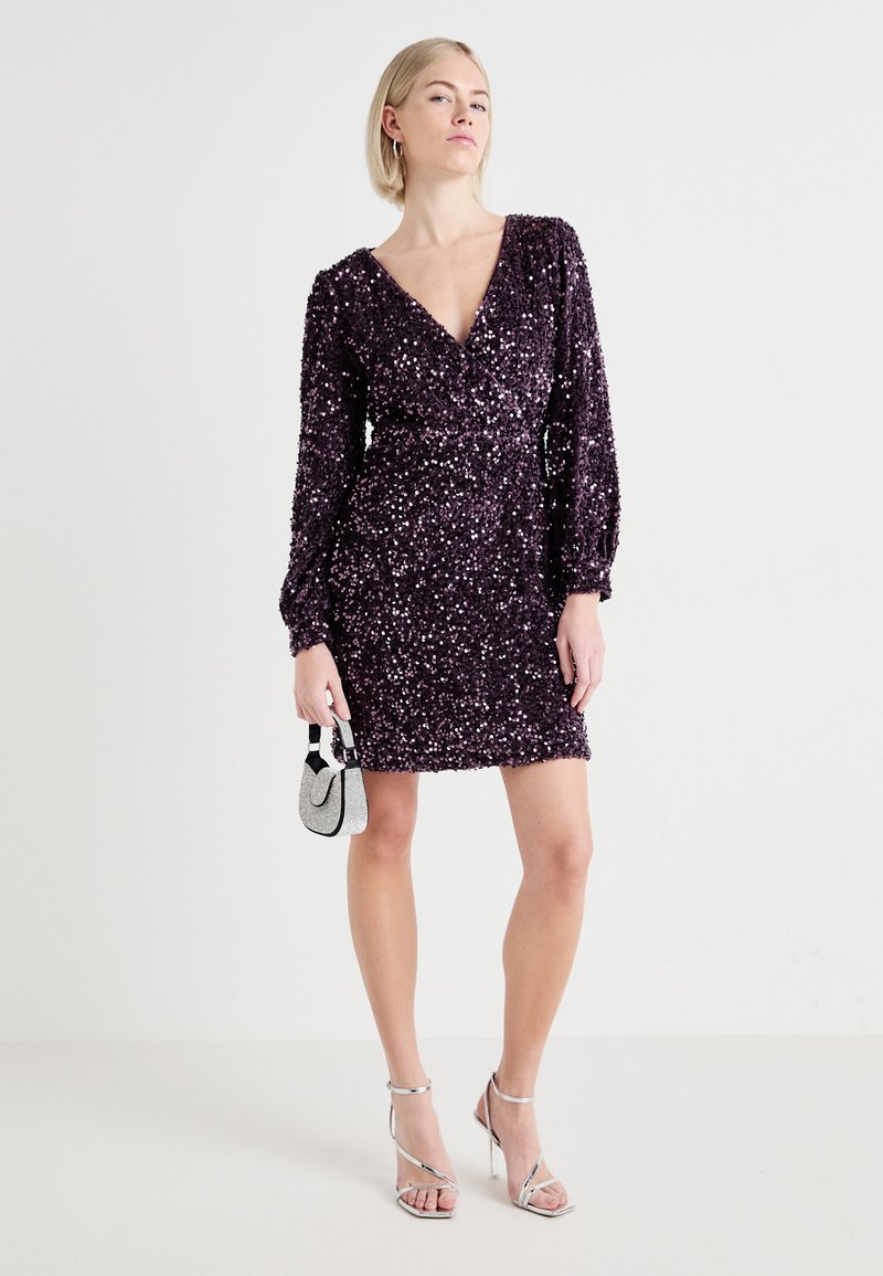 Krótka sukienka z purpurowymi cekinami, z głębokim dekoltem w kształcie V, długimi bufkami oraz przewiązaną talią. Połączona z srebrnymi sandałami na obcasie i małą torebką.