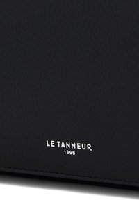 Producto de piel negra con una superficie texturizada, que presenta un logo plateado en relieve y la fecha "LE TANNEUR 1898" cerca del borde inferior.