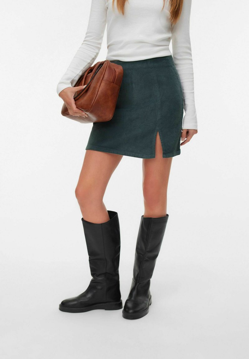 Mini-jupe en velours côtelé vert avec une fente sur le côté, associé à un top ajusté à manches longues blanc et un sac à main en cuir marron, complété par des bottes noires à hauteur de genoux.