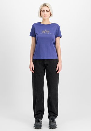Camiseta de manga corta morada con estampado de logo dorado, combinada con jeans sueltos negros y zapatillas negras. Textura de algodón suave.