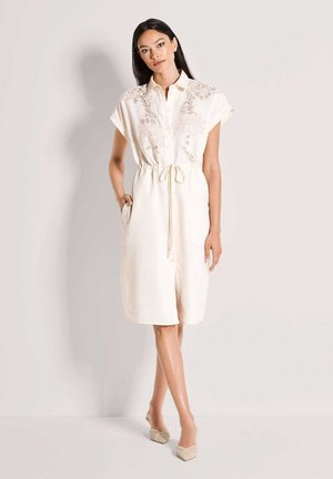 Crèmekleurige linnen jurk met korte mouwen, voorzien van een knoopsluiting aan de voorkant, bloemenkant bij de halslijn en een taille met trekkoord.