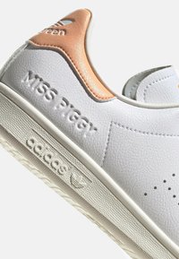 White leather sneaker with a textured orange accent on the heel, embossed "Miss Piggy" branding, and a rubber sole with a logo.