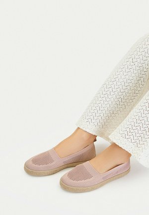 Piedi con scarpe slip-on in maglia rosa chiaro con suole beige intrecciate e pantaloni bianchi strutturati su sfondo bianco.