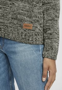 Grå och svart stickad tröja med ett strukturerat mönster, med en läderetikett märkta "OXMO" nära fållen, kombinerad med blå denimjeans.