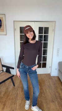 Noor naeratav naine, kes kannab pruunat pikkade varrukatega särki, siniseid teksaseid, pruuni nokamütsi ja valgeid tossusid, seisab toas, millel on puidust põrand ja valged uksed.