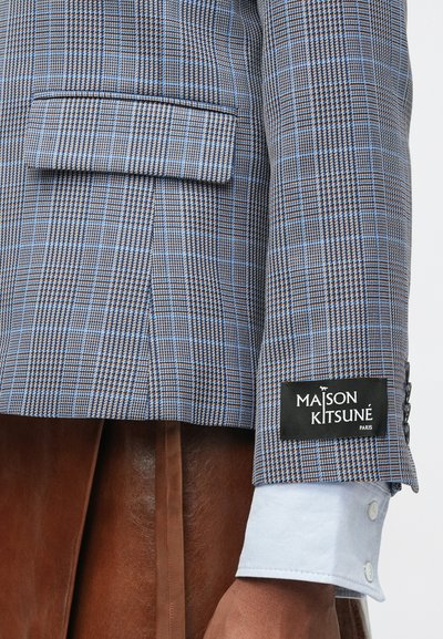 Kék és barna kockás blézer, mellzsebével és egy fekete címkével, amelyen a "MAISON KITSUNÉ PARIS" felirat található. Világoskék mandzsetta részlet jellemzi.