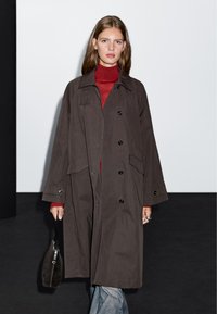 Długi, ciemnobrązowy płaszcz z zapięciem z przodu, dużymi kieszeniami i guzikami; nałożony na czerwony golf. Modelka trzyma małą torebkę.