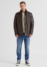 Braune Lederjacke mit Stehkragen, Reißverschlusstasche und Druckknopfverschlüssen, kombiniert mit hellblauen Jeans und schwarz-weißen Turnschuhen.